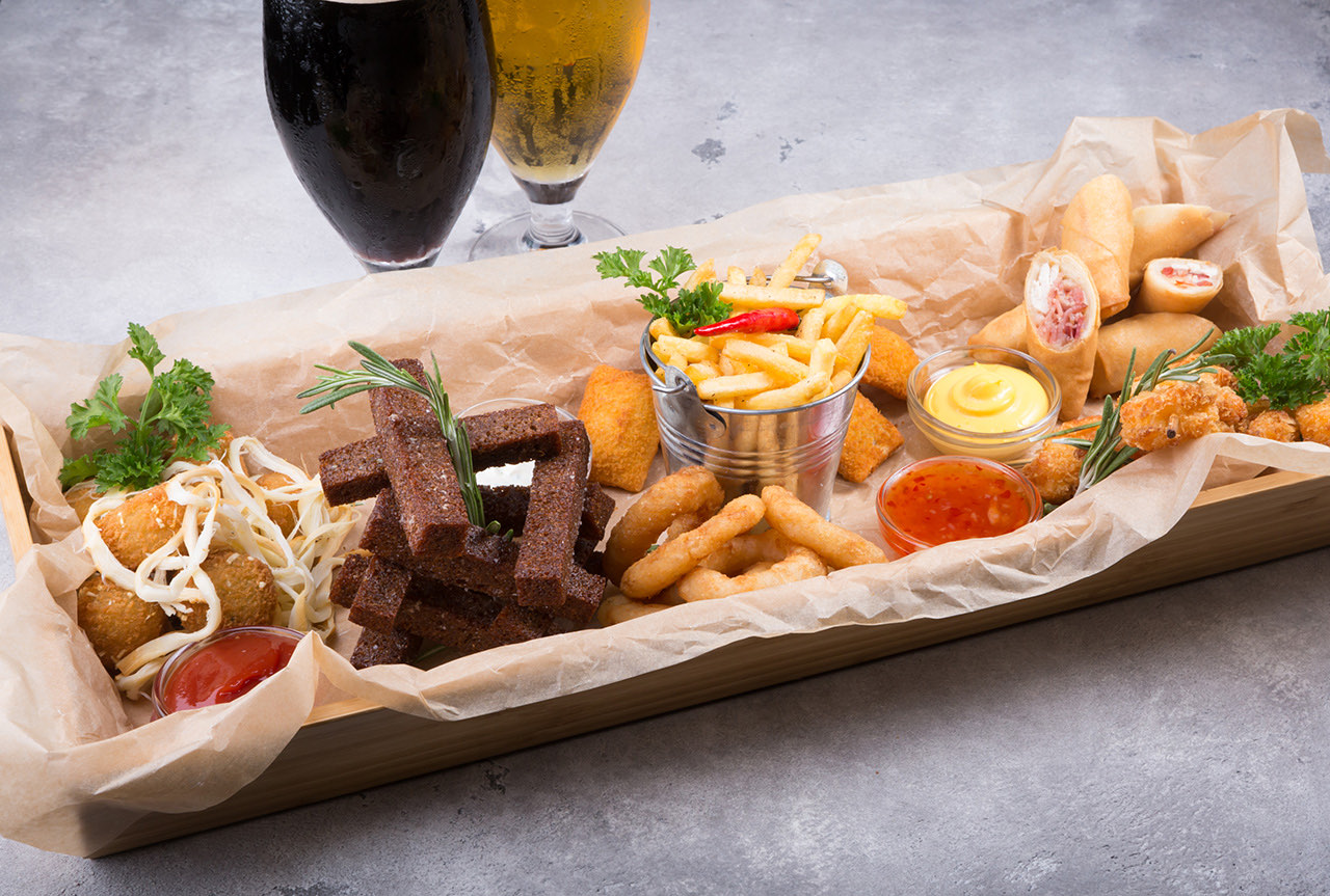 A large shareable appetizer platter featuring French fries, fried snacks, breadsticks, spring rolls and dipping sauces, served in a parchment-lined wooden tray.